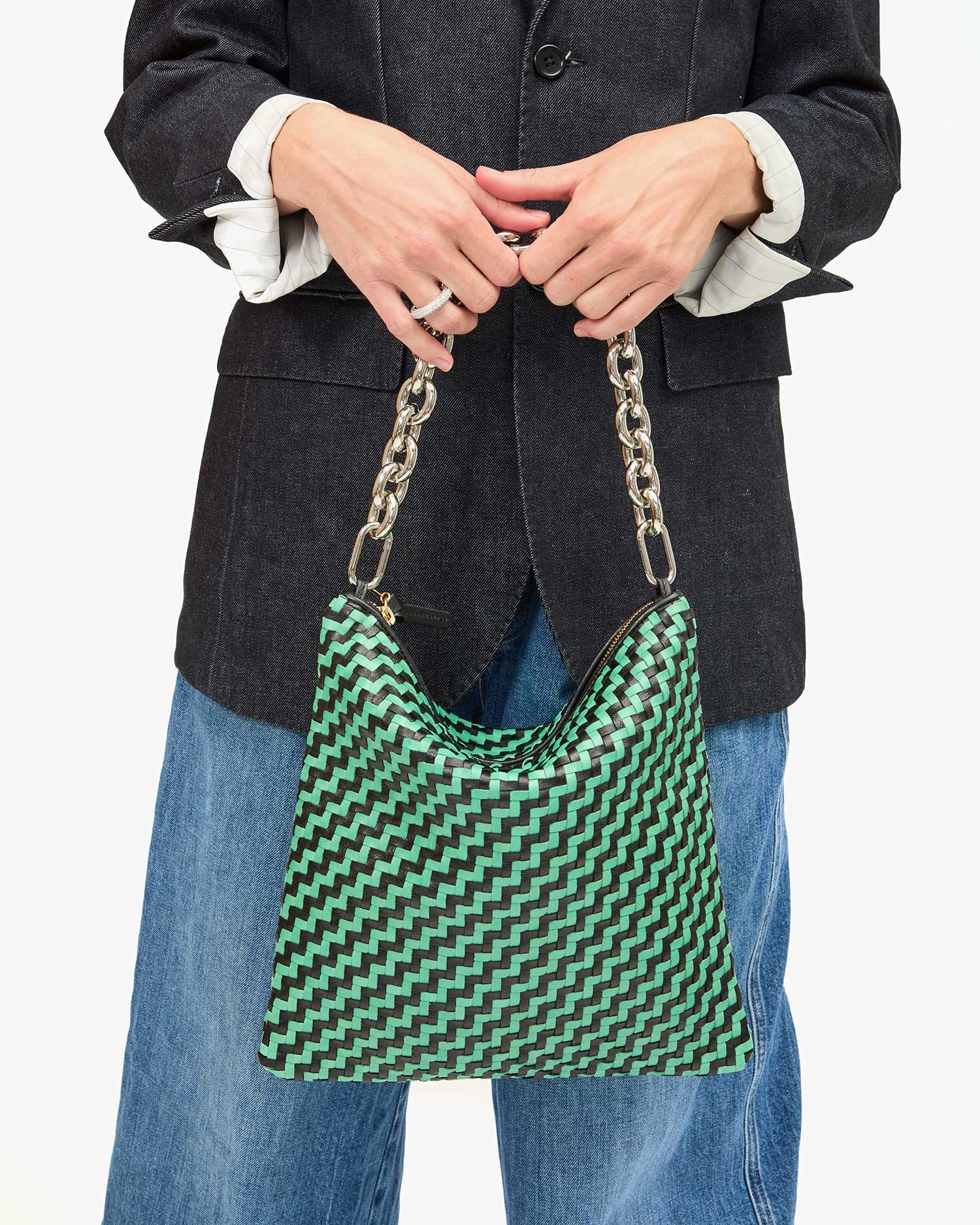 A person wearing a dark blazer and blue jeans holds the Foldover Clutch w/Tabs by Clare Vivier, featuring a stylish green and black pattern with a silver chain strap. The plain white background enhances the ensemble&#39;s elegance.