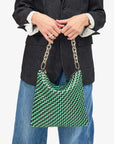 A person wearing a dark blazer and blue jeans holds the Foldover Clutch w/Tabs by Clare Vivier, featuring a stylish green and black pattern with a silver chain strap. The plain white background enhances the ensemble's elegance.