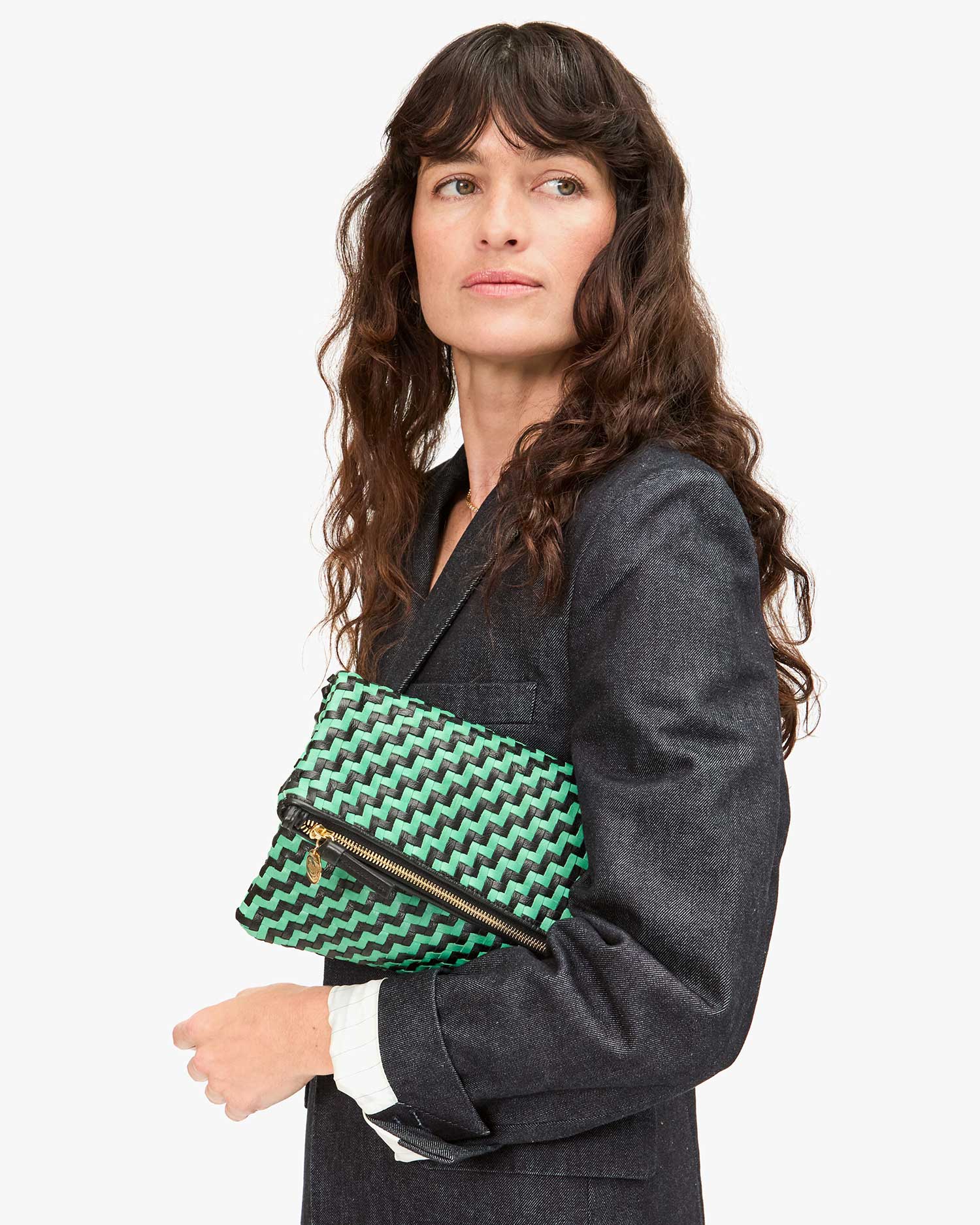 A person with long, wavy hair is looking to the side while holding a Foldover Clutch w/Tabs by Clare Vivier featuring a green zigzag pattern. They are dressed in a dark blazer over a white shirt.