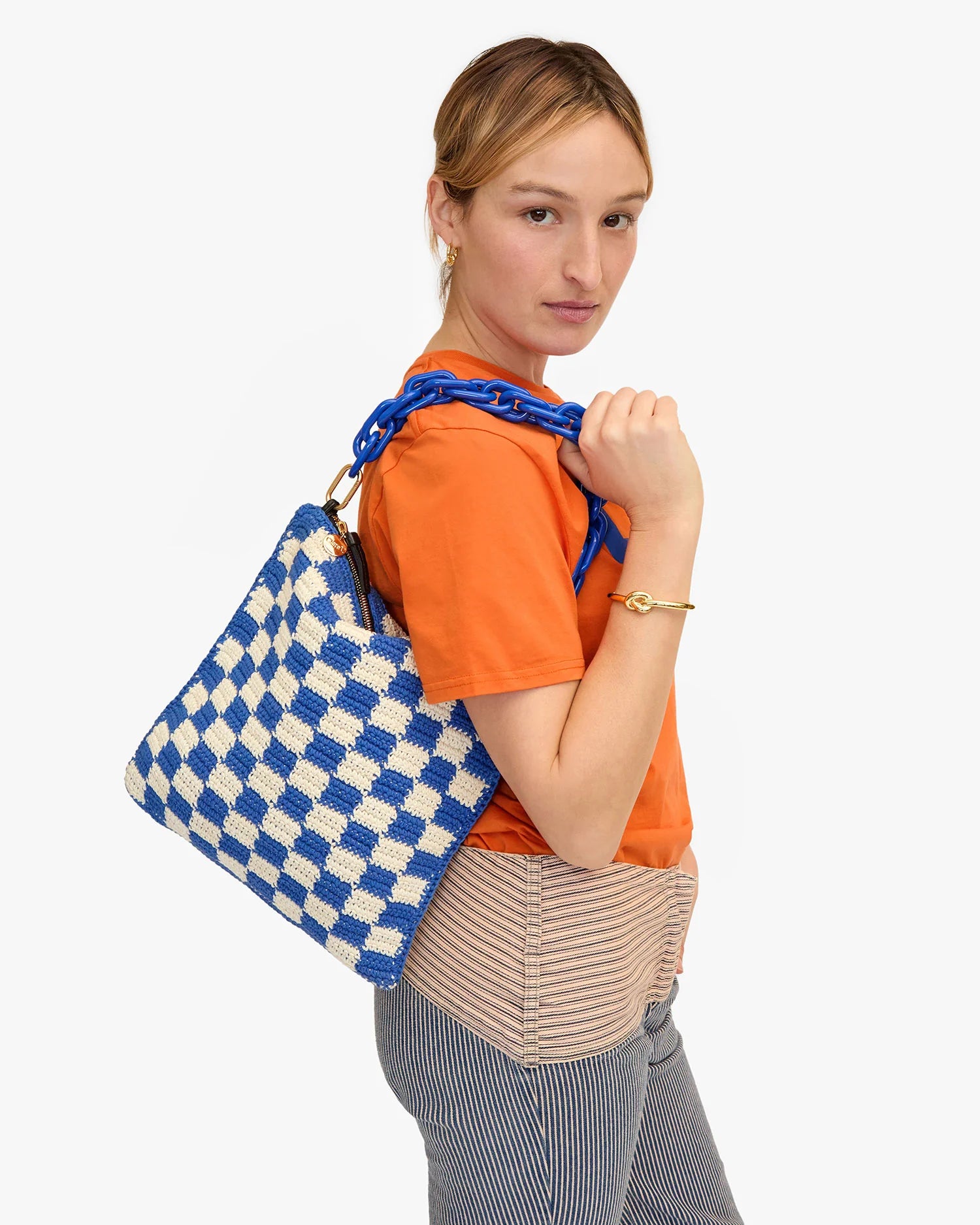 A person with light brown hair is shown from the waist up, wearing an orange t-shirt and striped pants. They are holding a Foldover Clutch w/Tabs by Clare Vivier with a blue chain strap over their shoulder. The bag, made of crocheted cotton, stands out vividly against the white background.