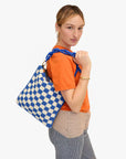 A person with light brown hair is shown from the waist up, wearing an orange t-shirt and striped pants. They are holding a Foldover Clutch w/Tabs by Clare Vivier with a blue chain strap over their shoulder. The bag, made of crocheted cotton, stands out vividly against the white background.