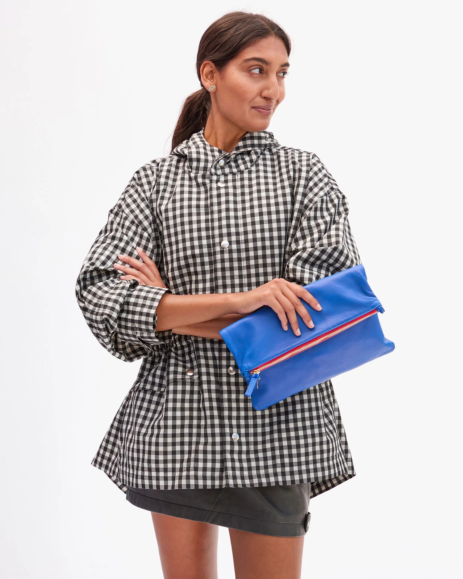 A woman in a black-and-white checkered jacket and dark skirt stands with arms crossed, holding the Clare Vivier Foldover Clutch W/ Tabs 26 in bright blue nappa leather, looking to the side against a plain white background.