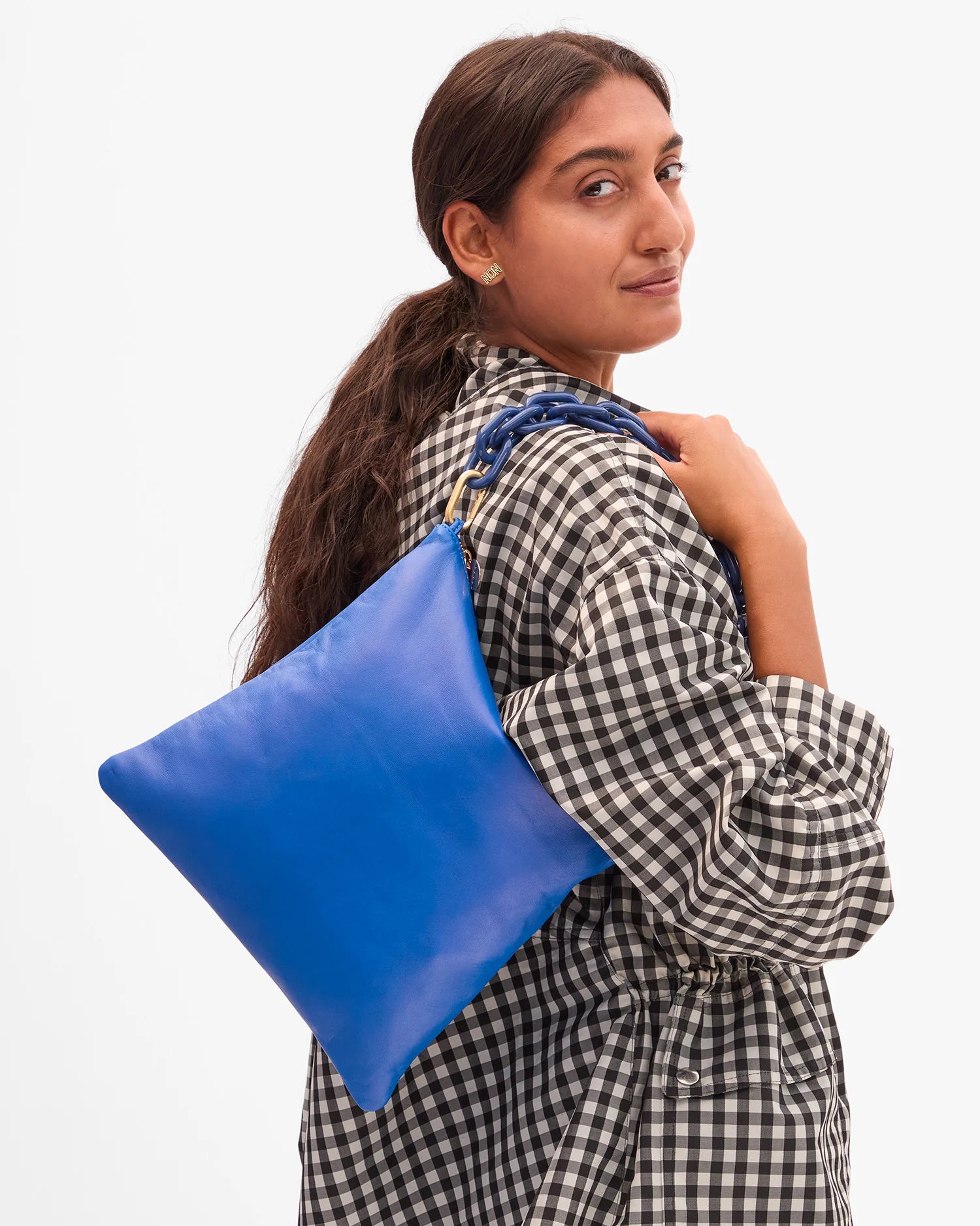 A woman in a black and white checkered shirt smiles over her shoulder, holding the bright blue Clare Vivier Foldover Clutch W/ Tabs 26 against a plain white background.