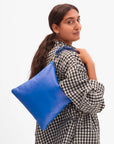 A woman in a black and white checkered shirt smiles over her shoulder, holding the bright blue Clare Vivier Foldover Clutch W/ Tabs 26 against a plain white background.