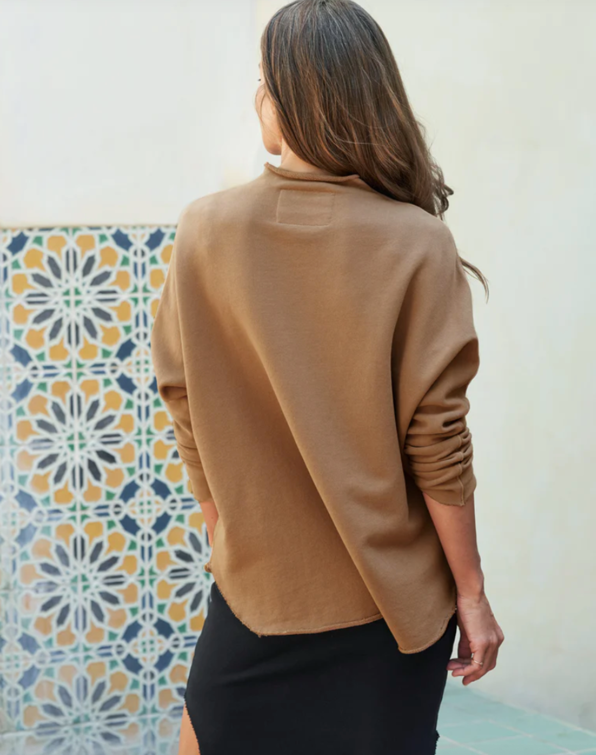 A woman with long brown hair stands near a geometric tile wall, wearing the Frank & Eileen EFFIE L/S Capelet in tan and a black skirt with a slit, her back facing the camera.