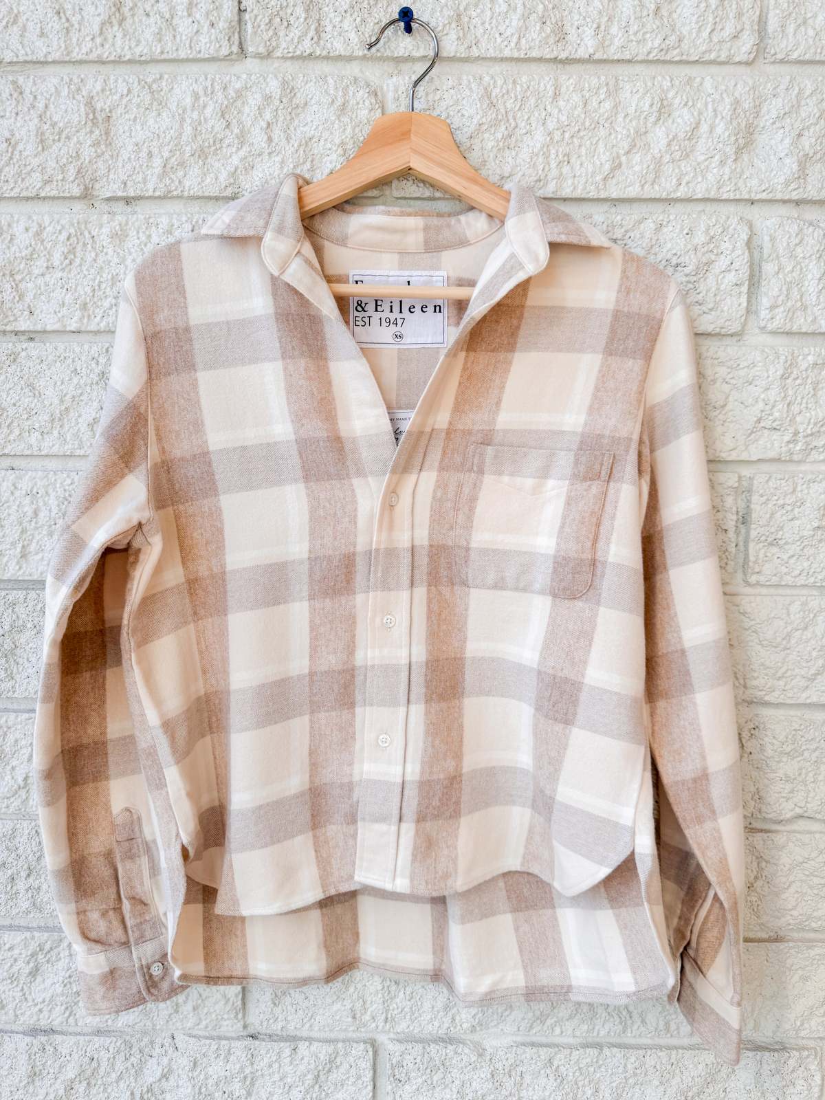 The Silvio Untuckable Shirt by Frank & Eileen, featuring a beige and white plaid pattern and collared design, hangs on a wooden hanger against a light brick wall.