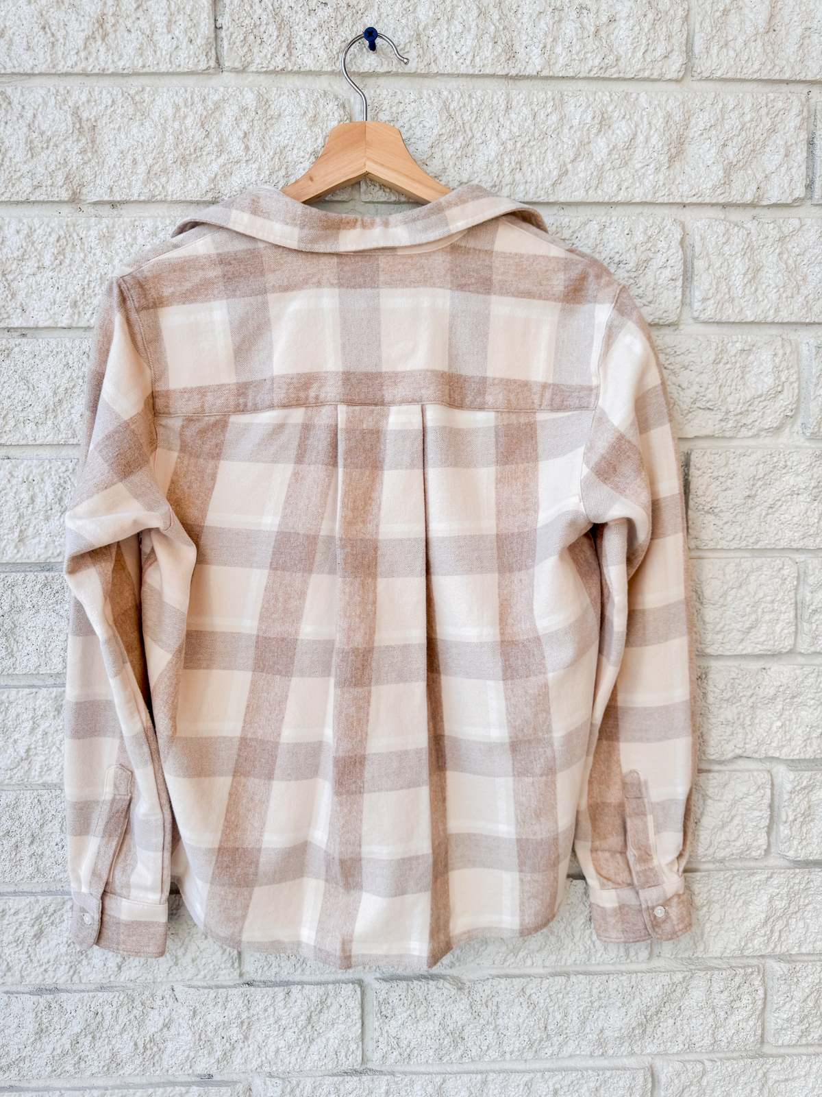 A Silvio Untuckable Shirt by Frank & Eileen in beige and light brown plaid hangs on a wooden hanger against a white brick wall, displayed from the back with sleeves down and collar open.