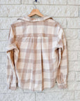 A Silvio Untuckable Shirt by Frank & Eileen in beige and light brown plaid hangs on a wooden hanger against a white brick wall, displayed from the back with sleeves down and collar open.