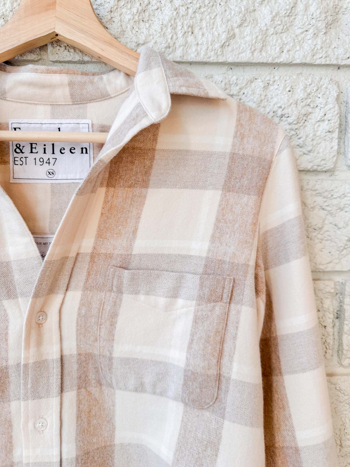 A cream and light brown plaid Silvio Untuckable Shirt by Frank & Eileen, featuring a chest pocket and a branded label, hangs on a wooden hanger against a white brick wall.