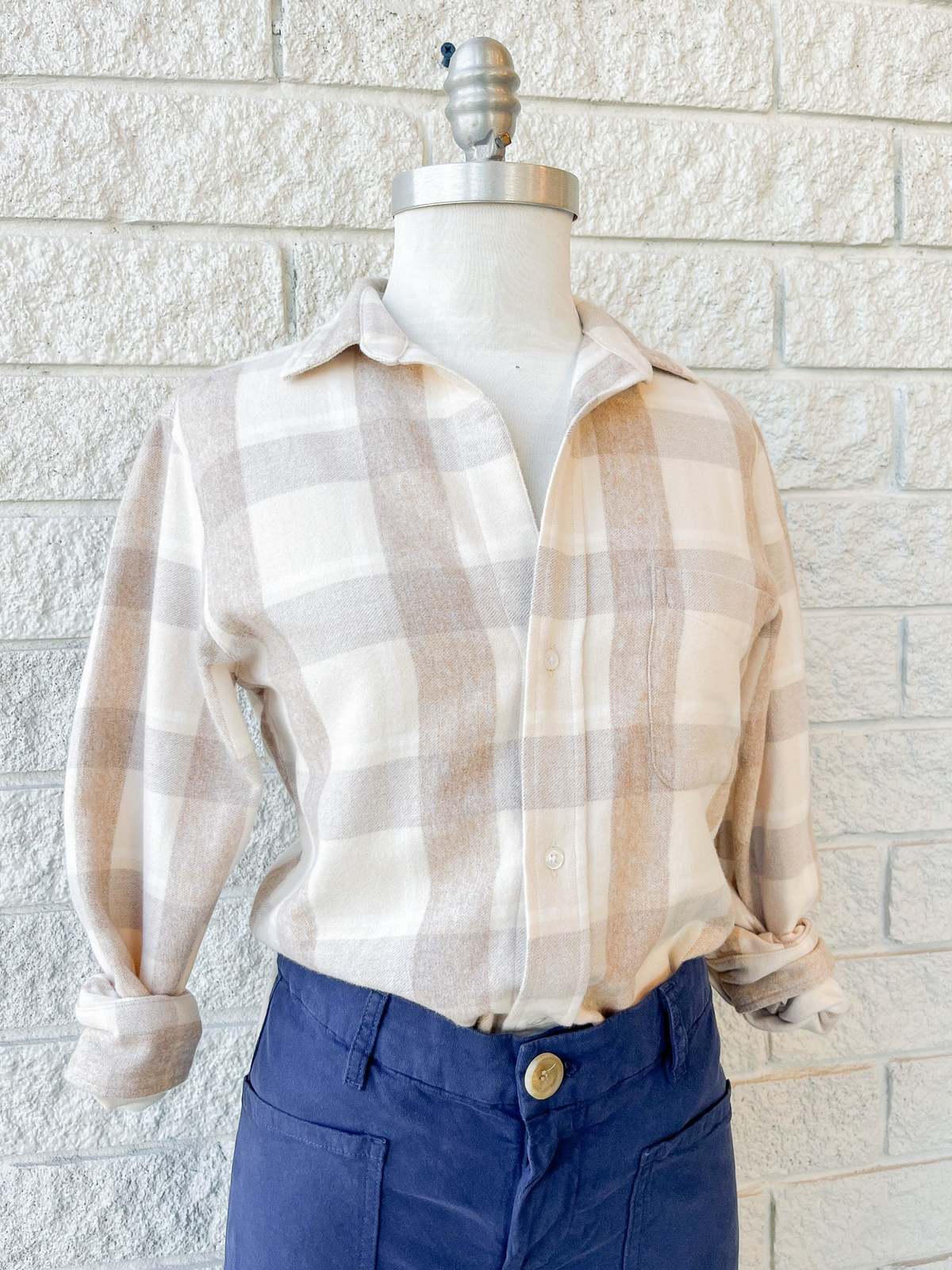 A mannequin wears Frank & Eileen's Silvio Untuckable Shirt in beige and cream plaid, featuring a petite square fit, paired with high-waisted navy pants, standing before a white brick wall.
