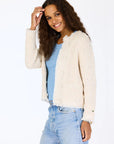 A woman with long curly hair smiles in a Kerri Rosenthal Fringe Beaulero Cardigan, layered over a light blue shirt and light-wash jeans, standing against a plain white background.
