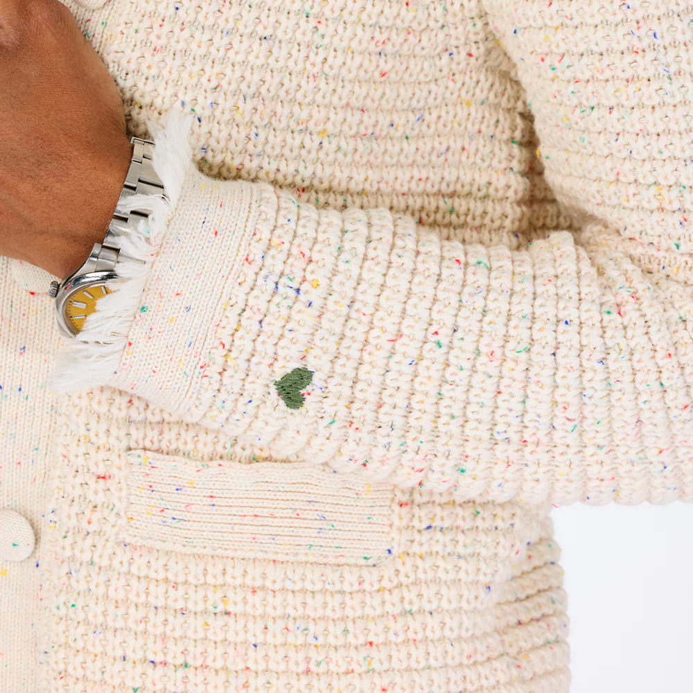 A person wears the Kerri Rosenthal Fringe Beaulero Cardigan, a cream-colored speckled knit with pockets. They also have a silver wristwatch on their left wrist, hand partially visible against a white background.