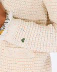A person wears the Kerri Rosenthal Fringe Beaulero Cardigan, a cream-colored speckled knit with pockets. They also have a silver wristwatch on their left wrist, hand partially visible against a white background.