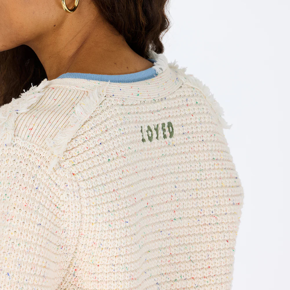 A woman wears the Kerri Rosenthal Fringe Beaulero Cardigan, a cream piece with colorful specks, frayed edges, and "LOVED" in green near the back neckline. Only her shoulder, neck, and part of her face are visible.