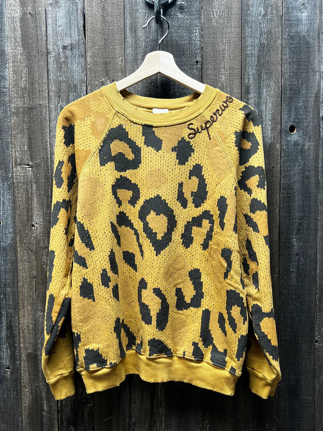 A "C&S Sweats Into the Wild Superwoman" sweater from the I Stole My Boyfriend's Shirt brand is hanging on a wooden hanger against a rustic wooden wall. This stylish piece features an eye-catching yellow and black leopard print, with "Supreme" embroidered in black on the collar for an exclusive touch.