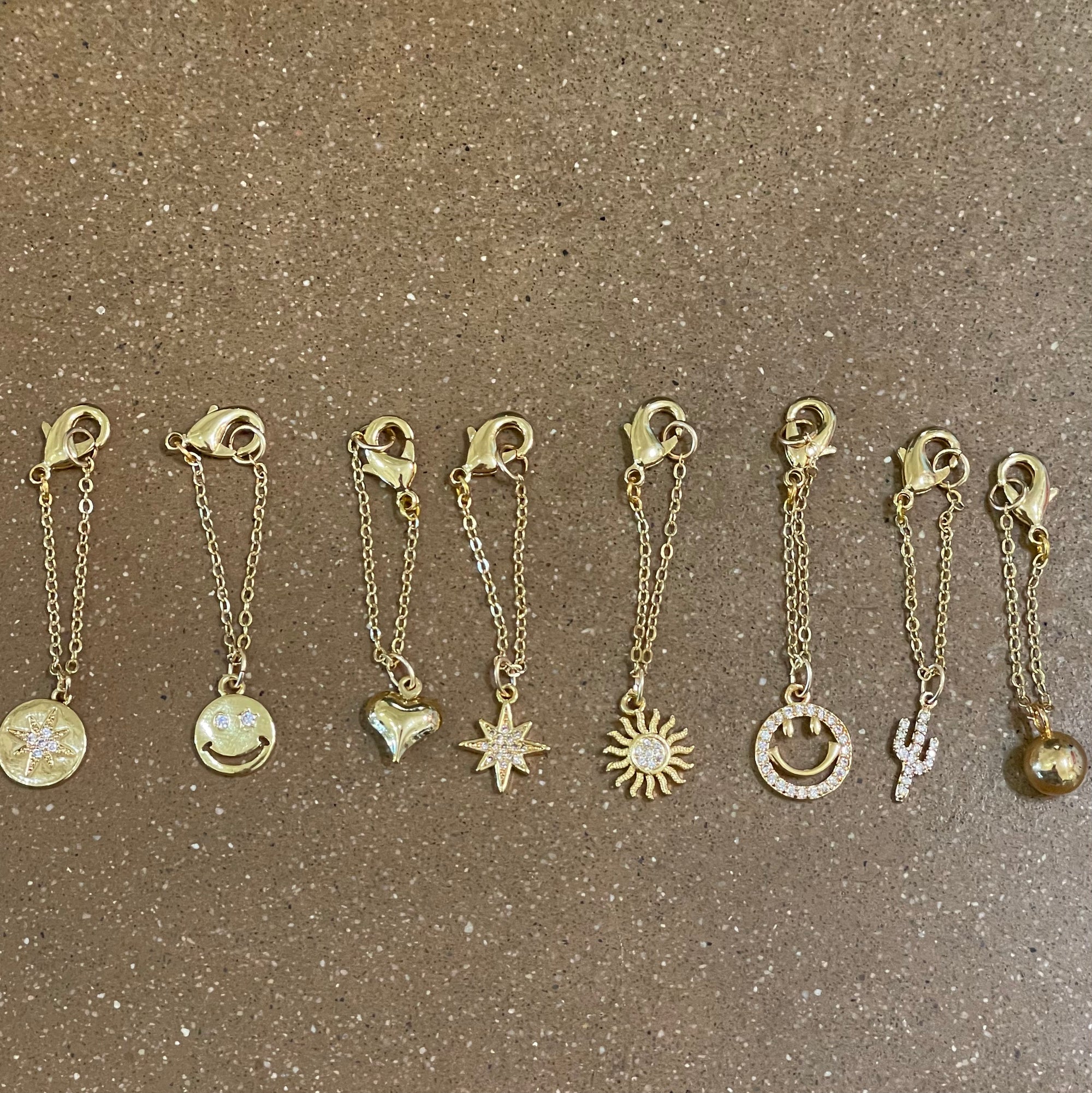 Seven Lucky Star Watch/Bag charms are arranged in a row on a textured brown surface. Each charm is distinct, showcasing designs such as a smiley face, heart, star, sun, crescent moon, cactus, and sphere.