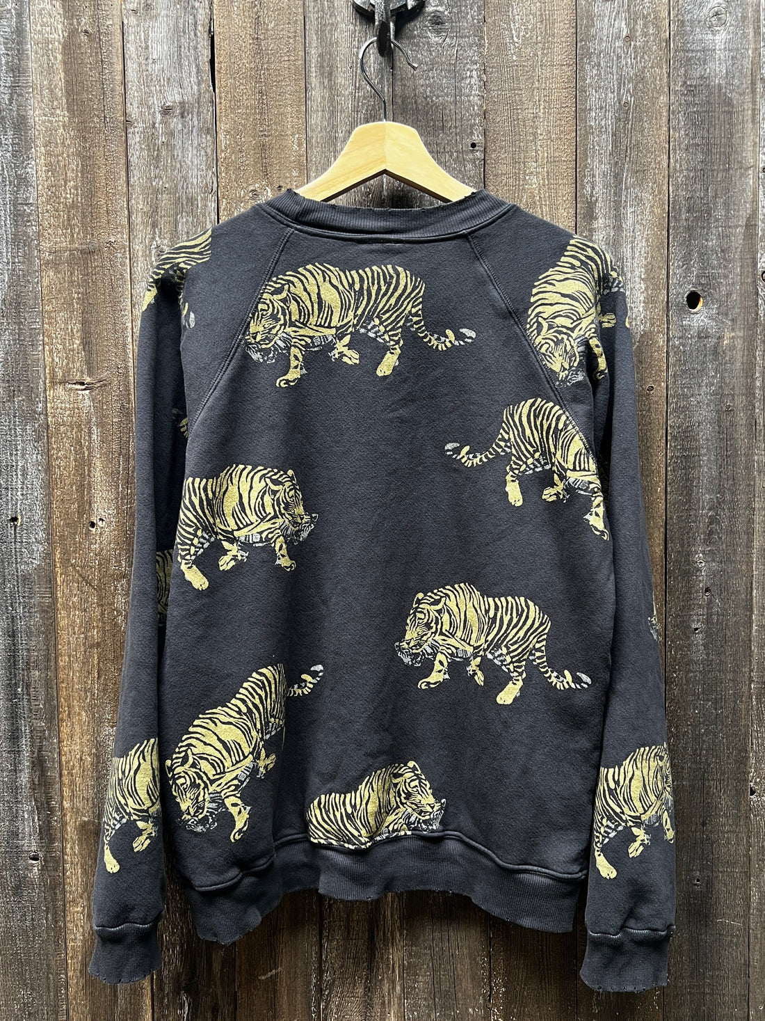 The "C&S Sweats Into the Wild Superwoman" from I Stole My Boyfriend's Shirt hangs on a wooden wall, featuring a silk screen-printed pattern of yellow tigers in various poses, with the wood background enhancing its rustic charm.