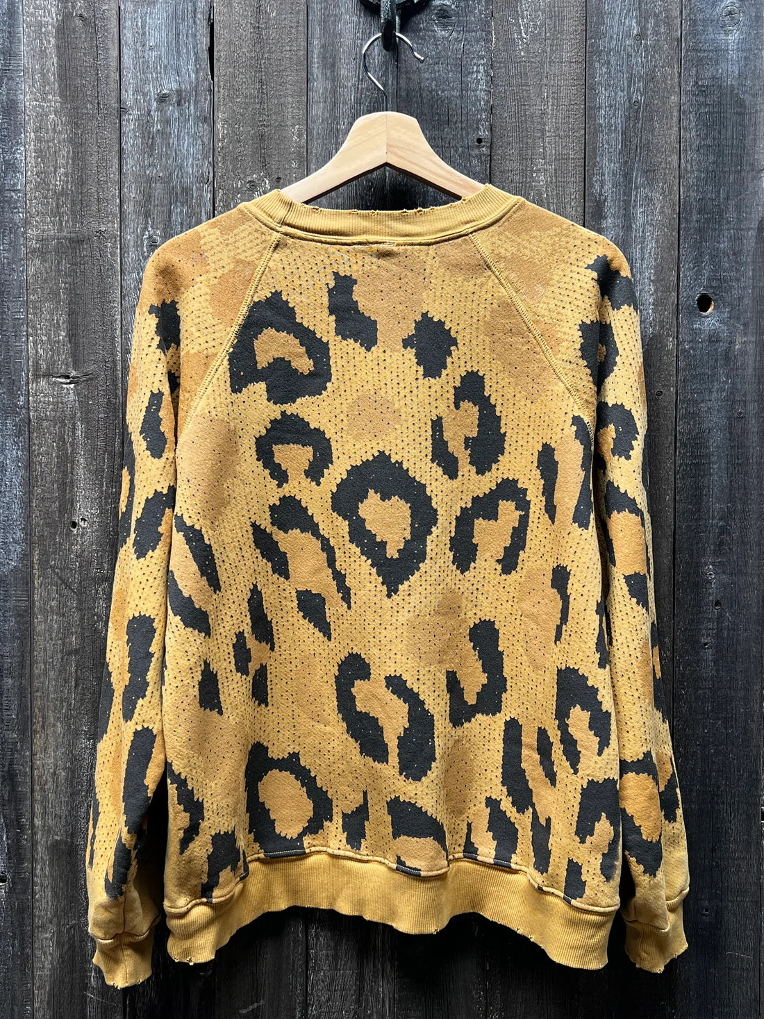 A "C&S Sweats Into the Wild Superwoman" sweater from I Stole My Boyfriend's Shirt hangs on a wooden hanger against a rough, dark wooden wall. This relaxed-fit, long-sleeve sweater presents an eye-catching animal pattern in yellow and black, combining elements of leopard and tiger for a bold fashion statement.