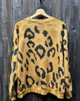 A "C&S Sweats Into the Wild Superwoman" sweater from I Stole My Boyfriend's Shirt hangs on a wooden hanger against a rough, dark wooden wall. This relaxed-fit, long-sleeve sweater presents an eye-catching animal pattern in yellow and black, combining elements of leopard and tiger for a bold fashion statement.