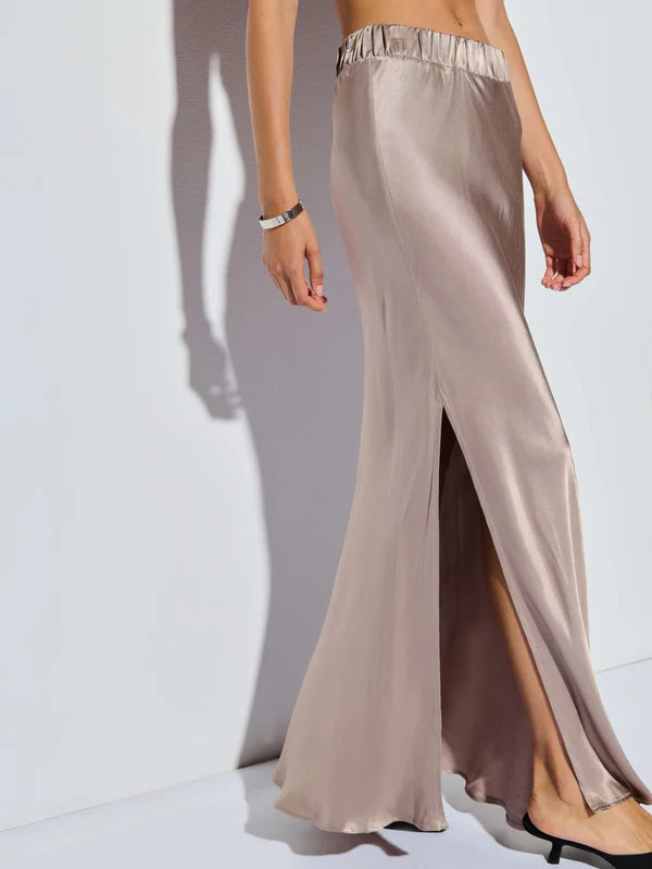 A person models the Maribel Bias Skirt w/ Slit by Nation LTD, crafted from champagne-hued hammered satin. It features an elastic waistband and a high slit, with its bias-cut hemline elegantly flowing and casting a shadow against the white backdrop.