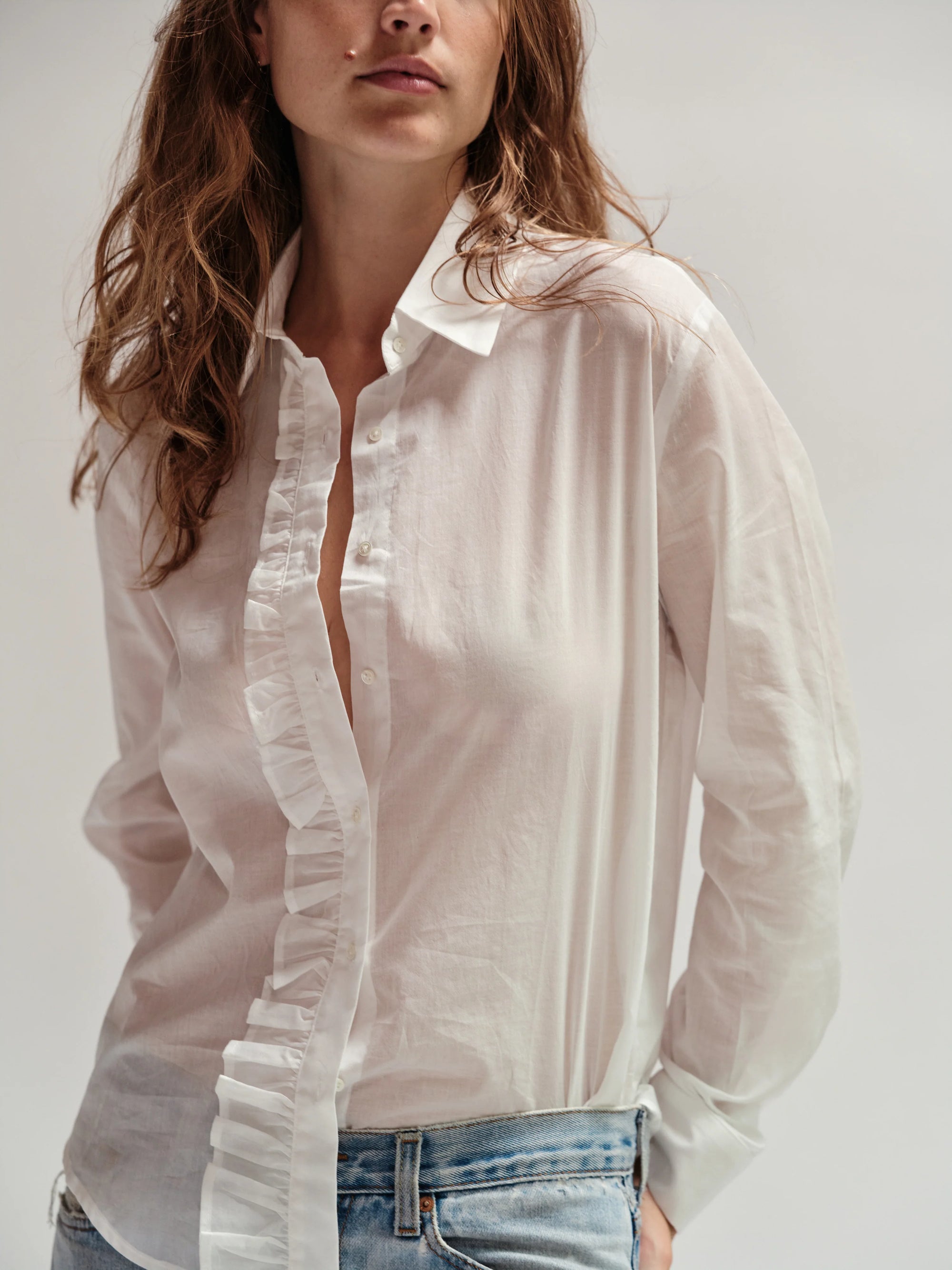 A woman with wavy brown hair wears Cissa’s The Waterfall Shirt, a sheer cotton ruffle top, paired with light blue jeans against a plain light background.