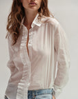 A woman with wavy brown hair wears Cissa’s The Waterfall Shirt, a sheer cotton ruffle top, paired with light blue jeans against a plain light background.