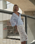 A woman stands by a metal fence in an urban setting, wearing Cissa's The Summer Pencil Skirt, a white embroidered piece with floral patterns, paired with a light blue oversized off-the-shoulder shirt.