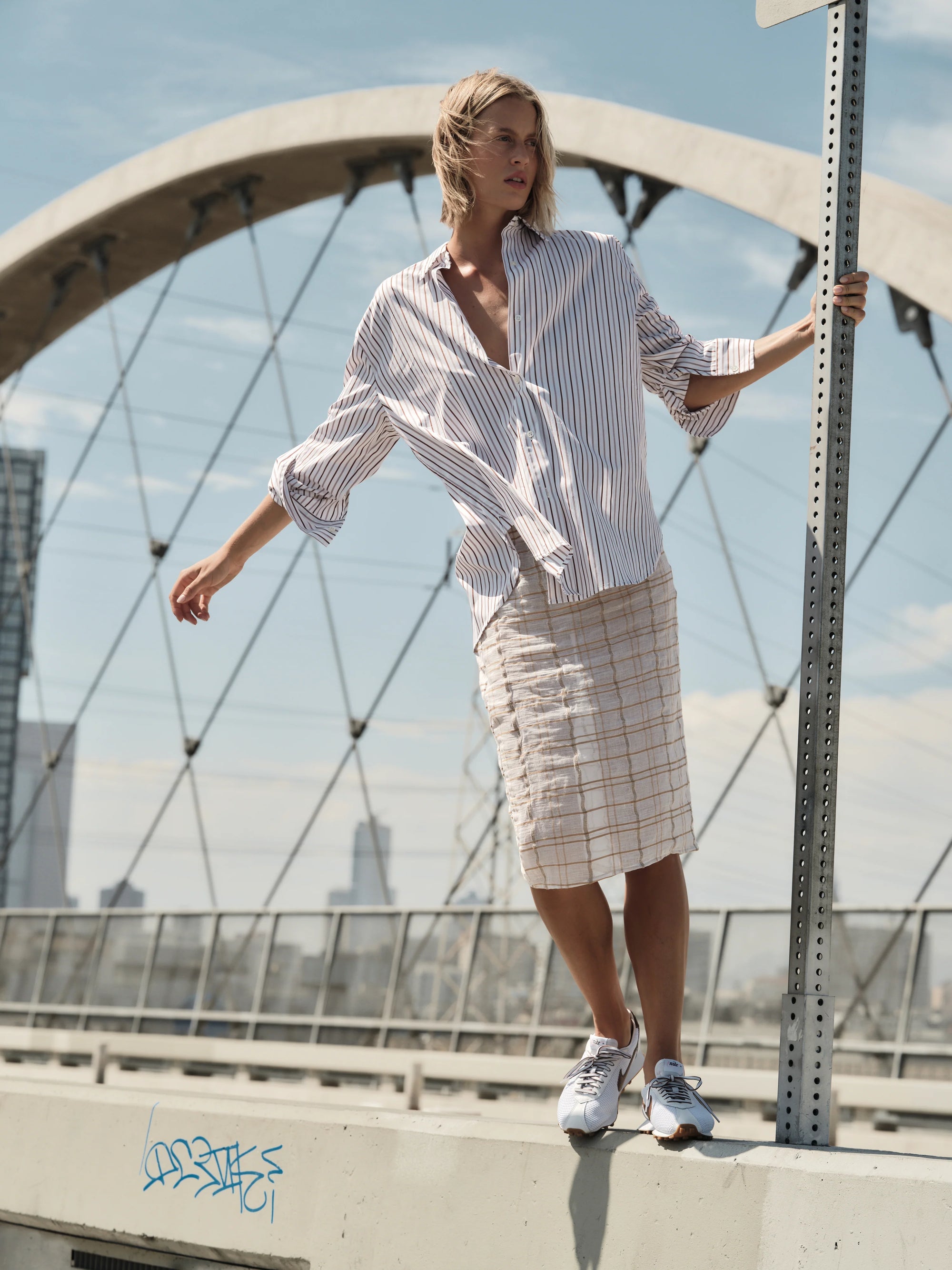 A person with short blonde hair and a striped shirt balances on a ledge near arched bridge supports, wearing the Cissa Summer Pencil Skirt and sneakers, with city buildings in the background on a sunny day.