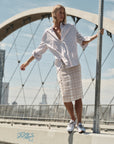 A person with short blonde hair and a striped shirt balances on a ledge near arched bridge supports, wearing the Cissa Summer Pencil Skirt and sneakers, with city buildings in the background on a sunny day.