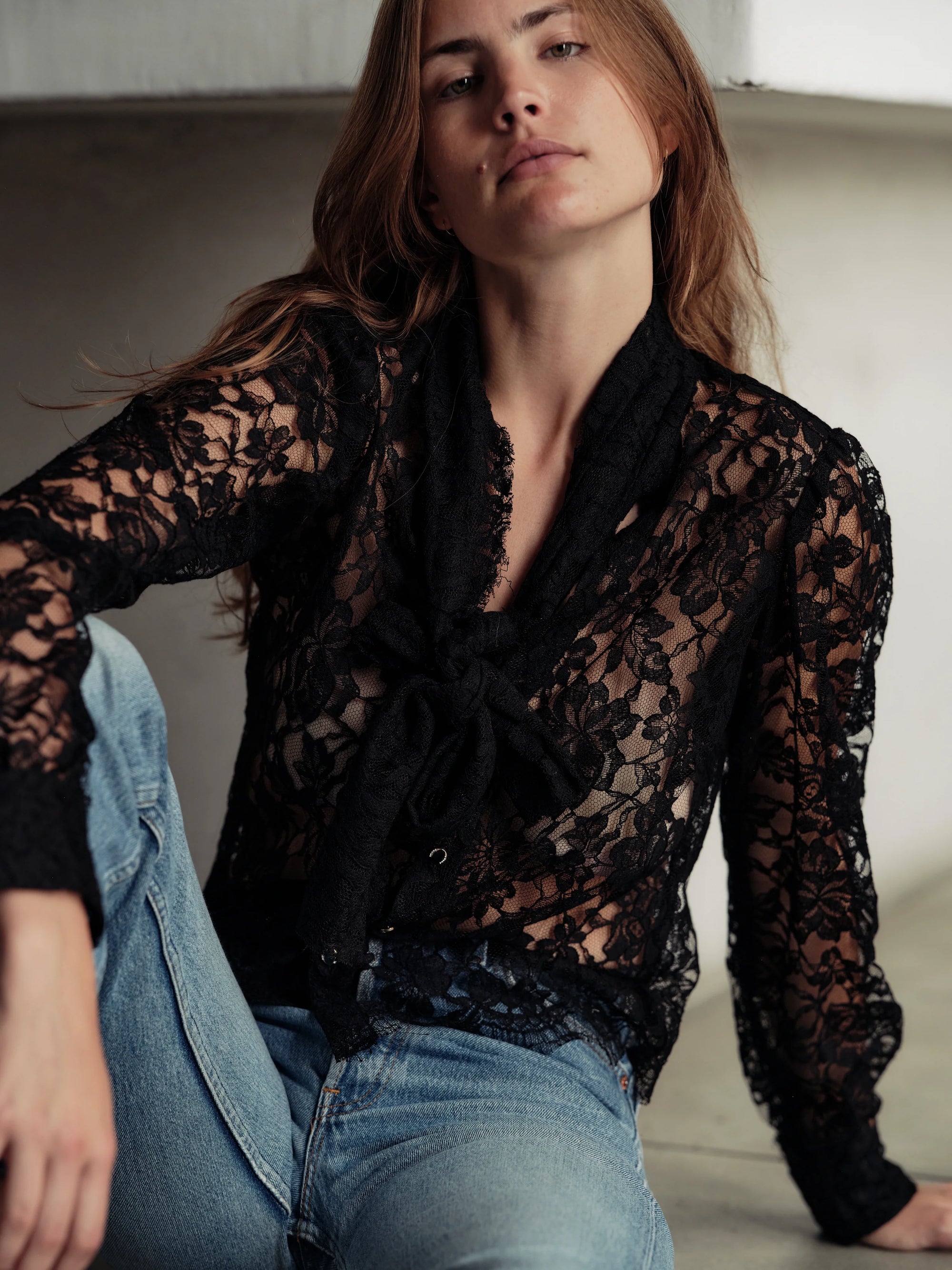 A woman with long brown hair models Cissa's "The Bow Tie Shirt," a sheer black lace blouse with a scalloped hemline, paired with blue jeans. She sits on the floor and looks at the camera with a relaxed expression.