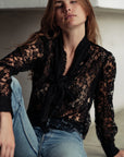 A woman with long brown hair models Cissa's "The Bow Tie Shirt," a sheer black lace blouse with a scalloped hemline, paired with blue jeans. She sits on the floor and looks at the camera with a relaxed expression.