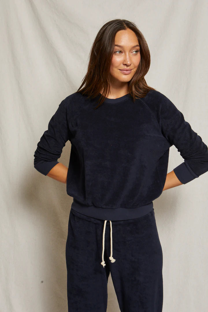 A woman with straight, center-parted brown hair stands against a light beige backdrop, wearing the Perfectwhitetee Saylor Loop Terry Sweatshirt in dark navy with matching drawstring pants. Her hands rest on her hips.
