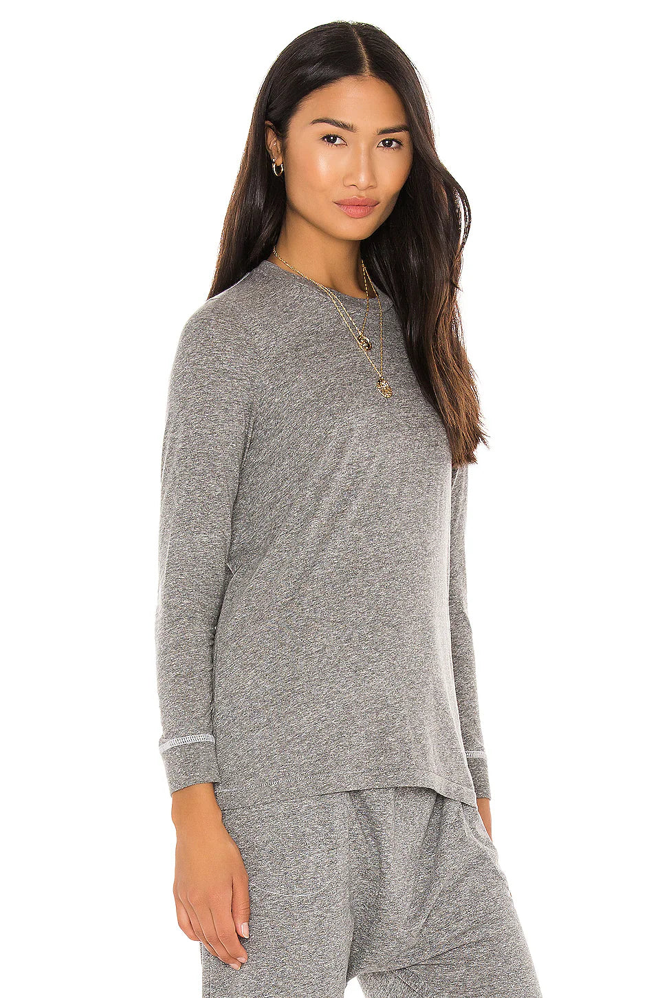 A woman with long, dark hair and gold jewelry smiles slightly against a white background, wearing The Great Inc.'s The LS Slim Tee—a grey cotton jersey long-sleeve top—paired with matching pants.