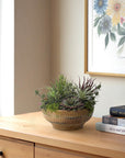The Succulent Wood Bowl by Natural Decorations Inc. sits on a light wooden dresser near books and floral artwork, creating a charming, maintenance-free display against a cream-colored wall by the window.