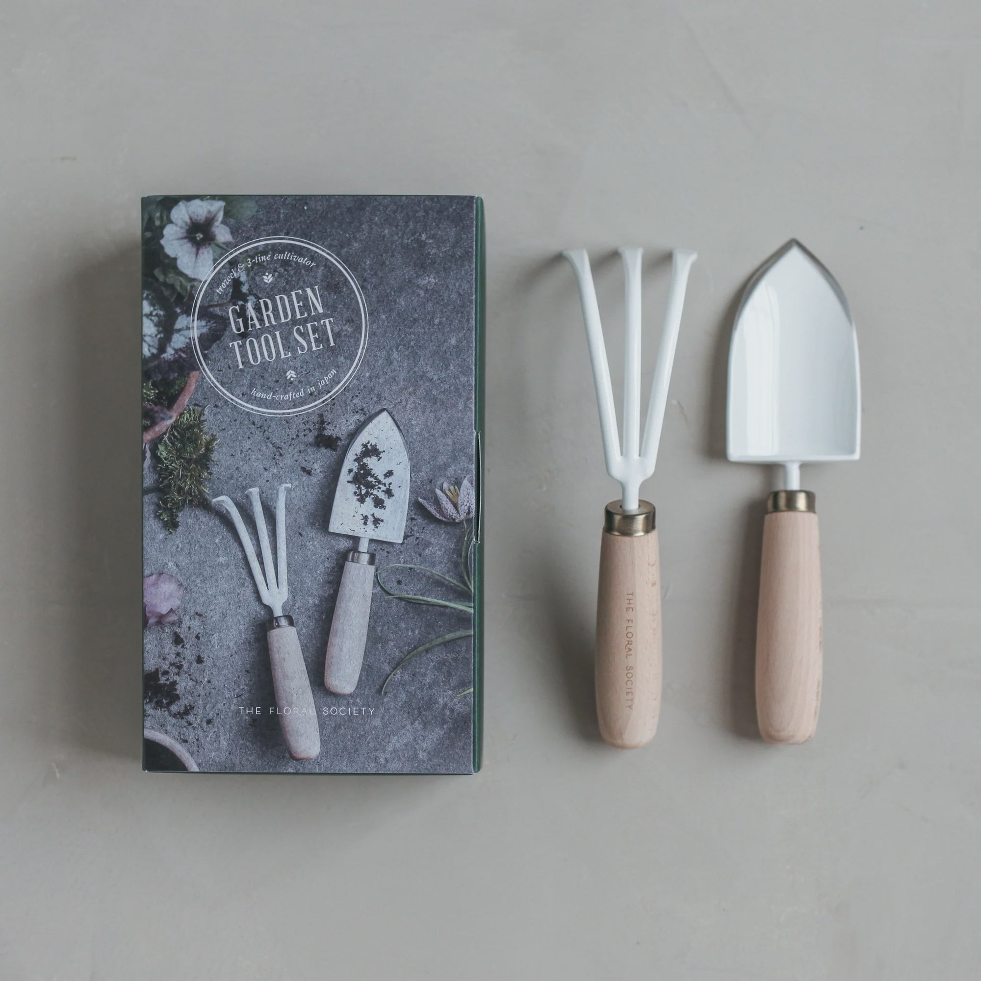 On a gray surface, the Garden Tool Set by The Floral Society is presented with hand-crafted mini rake and spade featuring white heads and wooden handles. The packaging displays images of the tools, including a detailed cultivator, set against vibrant flowers.