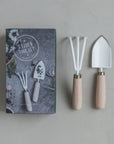 On a gray surface, the Garden Tool Set by The Floral Society is presented with hand-crafted mini rake and spade featuring white heads and wooden handles. The packaging displays images of the tools, including a detailed cultivator, set against vibrant flowers.