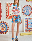 A woman in a room with colorful art on the walls wears a white sweater and a Hunter Bell NYC Powell Skirt in an Orange Grove print. Her blue, ankle-tied sandals grace the white floor, where a yellow tube lies.