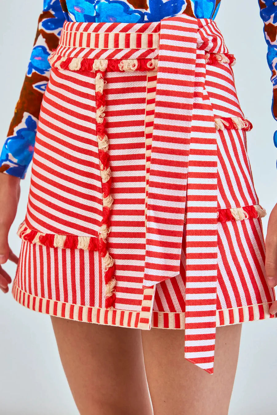 A person is seen in a close-up wearing the Bay Skirt by Hunter Bell NYC, featuring red and white yarn-dyed stripes with ruffled edges and a tied belt. The partially visible top showcases a bold blue floral pattern, making this ensemble truly vibrant.