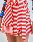 A person is seen in a close-up wearing the Bay Skirt by Hunter Bell NYC, featuring red and white yarn-dyed stripes with ruffled edges and a tied belt. The partially visible top showcases a bold blue floral pattern, making this ensemble truly vibrant.