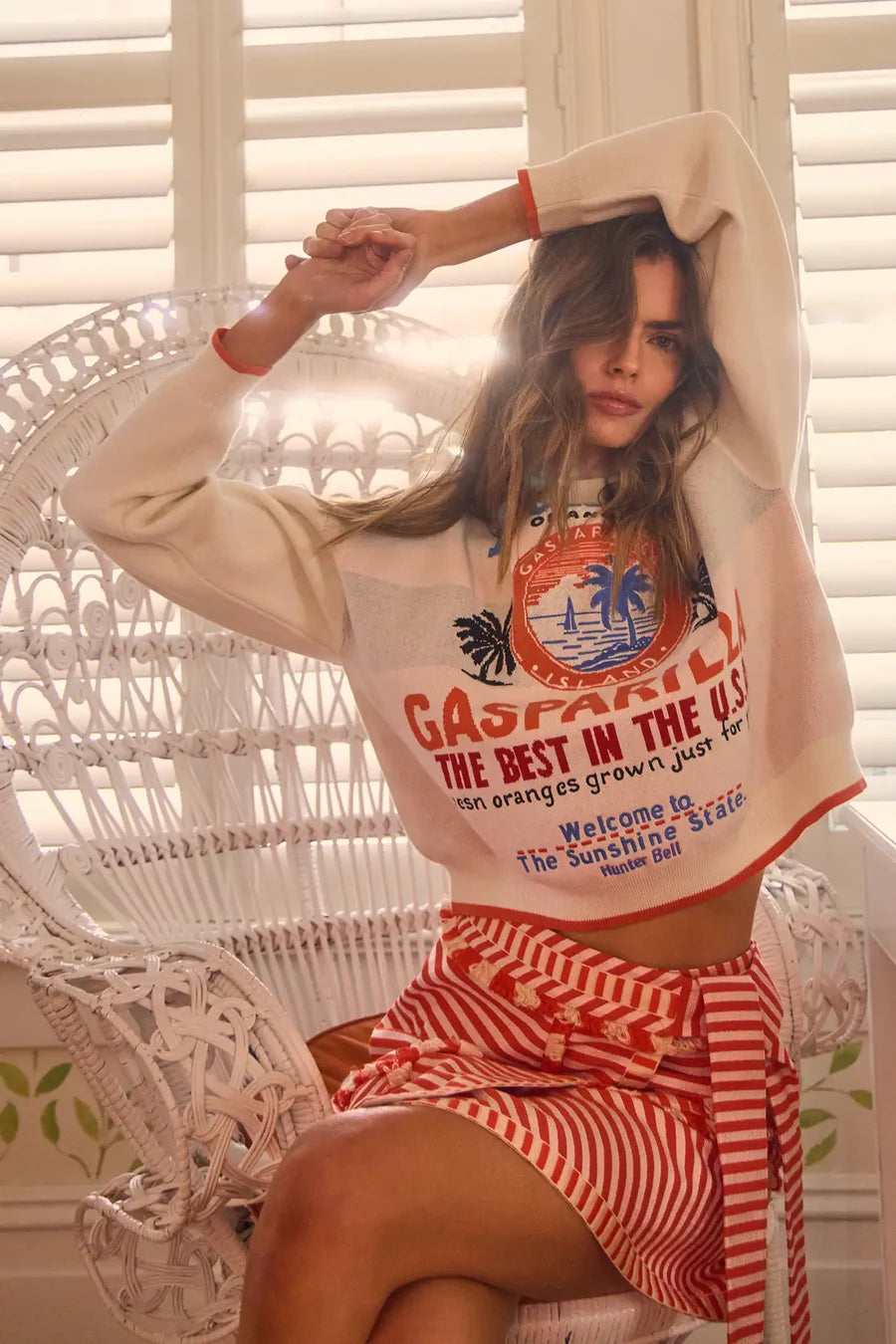 A woman poses indoors on a wicker chair, wearing a cream graphic sweater paired with Hunter Bell NYC's Bay Skirt, featuring red-and-white yarn-dyed stripes. Her long hair is loose, one arm raised against white shutters.