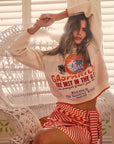 A woman poses indoors on a wicker chair, wearing a cream graphic sweater paired with Hunter Bell NYC's Bay Skirt, featuring red-and-white yarn-dyed stripes. Her long hair is loose, one arm raised against white shutters.