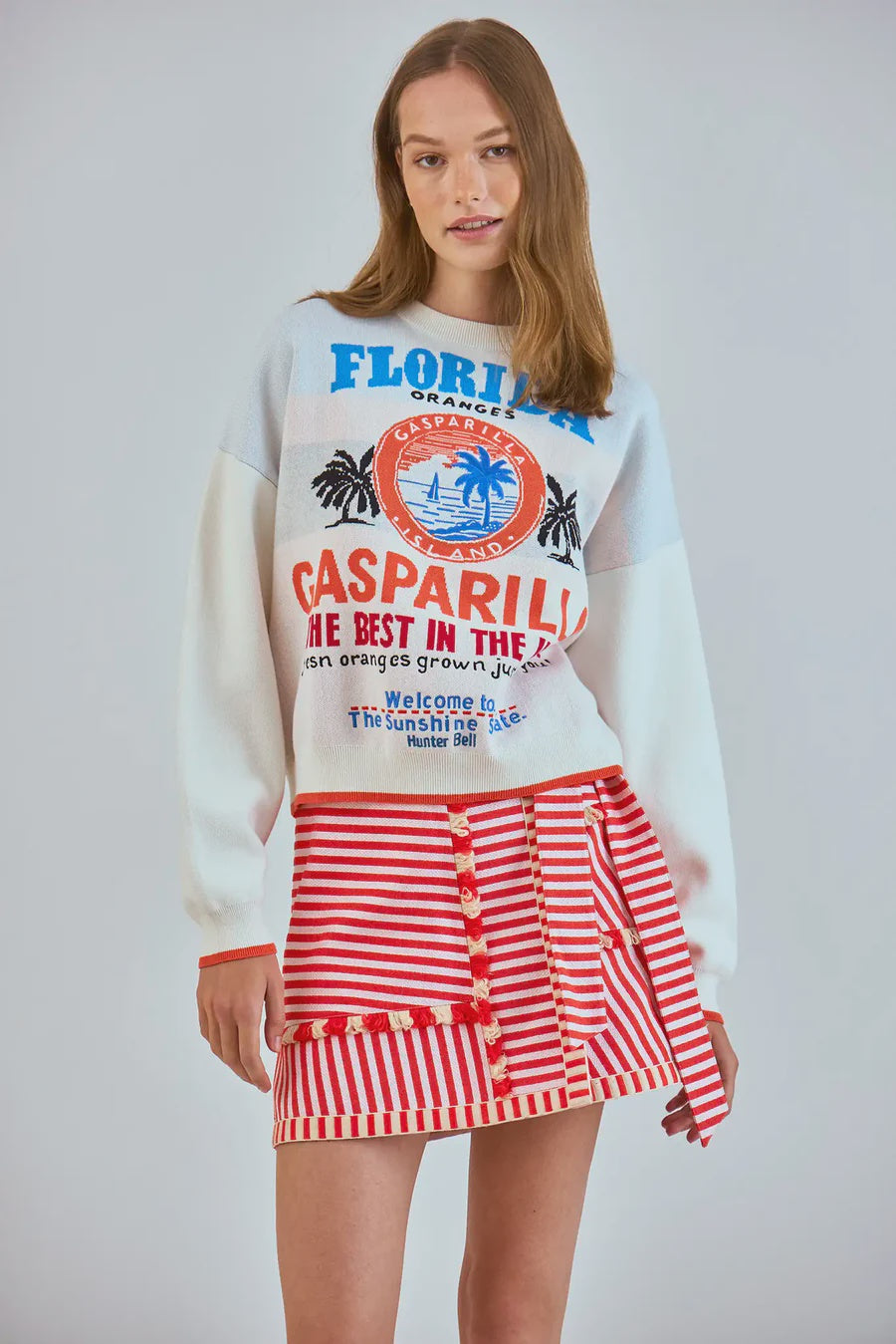 Wearing a white sweater featuring colorful text and graphics referencing Florida, Gasparilla, and oranges, a woman confidently models a red and white yarn-dyed striped Bay Skirt by Hunter Bell NYC against a neutral backdrop, gazing directly at the camera.