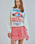 Wearing a white sweater featuring colorful text and graphics referencing Florida, Gasparilla, and oranges, a woman confidently models a red and white yarn-dyed striped Bay Skirt by Hunter Bell NYC against a neutral backdrop, gazing directly at the camera.