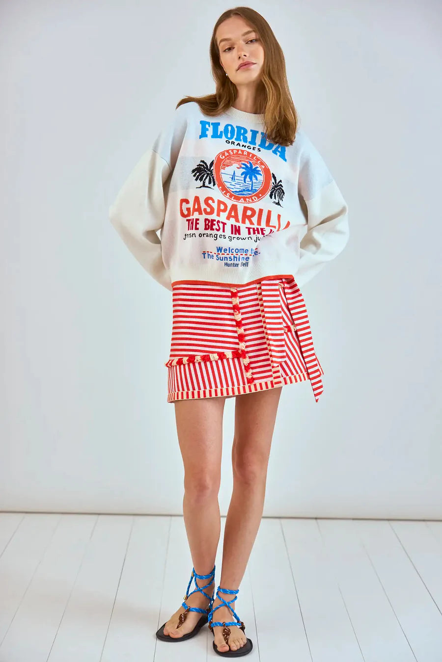 A woman wears a white sweatshirt with blue "Florida" text and palm trees, paired with Hunter Bell NYC's Bay Skirt in red and white yarn-dyed stripes. Blue lace-up sandals complete her look as she stands stylishly against a light wall on a pristine white floor.