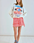 A woman wears a white sweatshirt with blue "Florida" text and palm trees, paired with Hunter Bell NYC's Bay Skirt in red and white yarn-dyed stripes. Blue lace-up sandals complete her look as she stands stylishly against a light wall on a pristine white floor.