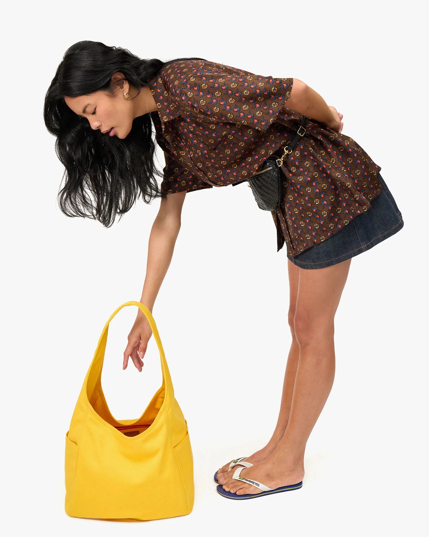 A woman with long dark hair, dressed in a patterned brown blouse, denim skirt, and flip-flops, bends forward to reach into a Clare Vivier Grand Bando Canvas tote bag resting on the floor.