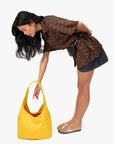 A woman with long dark hair, dressed in a patterned brown blouse, denim skirt, and flip-flops, bends forward to reach into a Clare Vivier Grand Bando Canvas tote bag resting on the floor.