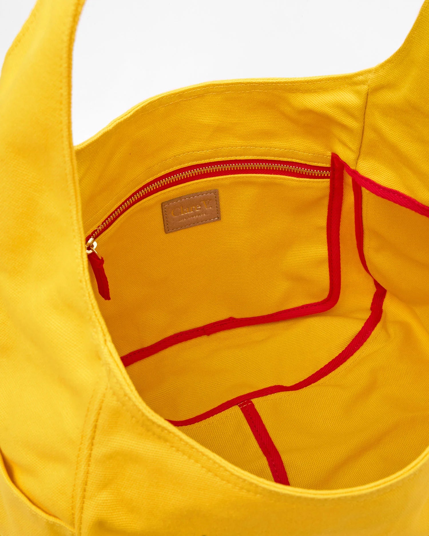 A close-up of the Grand Bando Canvas by Clare Vivier shows the yellow cotton tote’s interior, featuring a zippered pocket and red trim along the edges of its pockets.