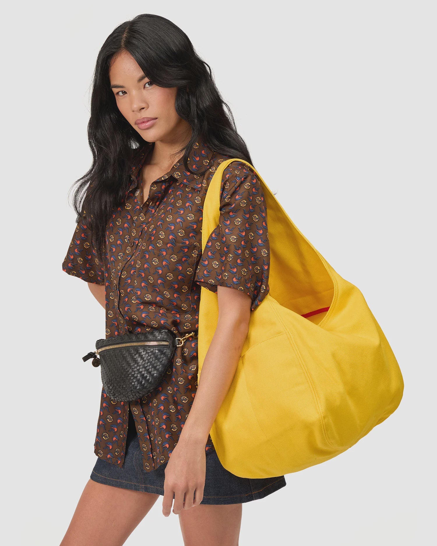 A woman with long black hair wears a brown patterned shirt, black skirt, and black woven belt bag. She carries Clare Vivier’s Grand Bando Canvas tote in bright yellow with external pockets against a plain light background.