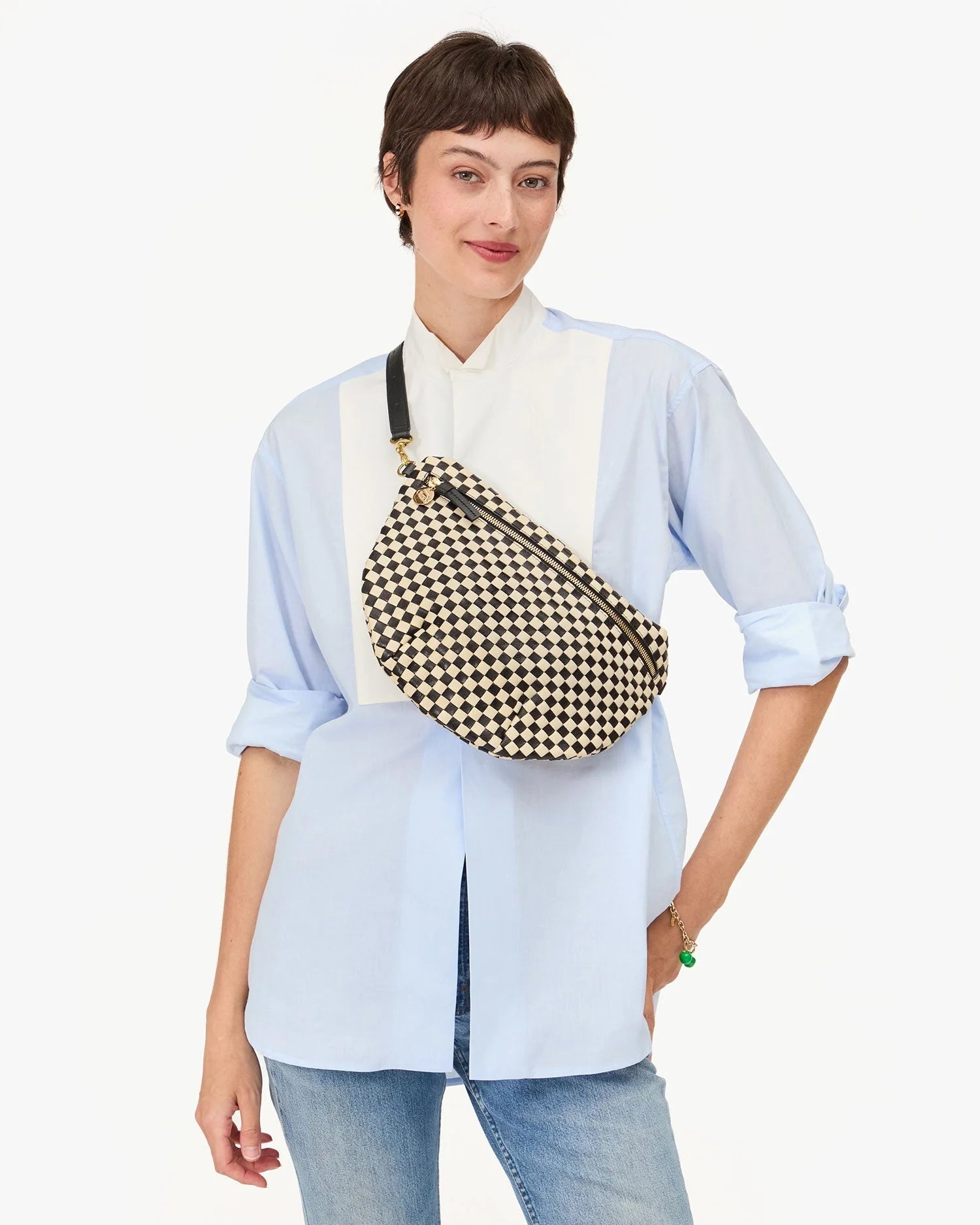 A smiling person in a light blue button-up shirt and jeans carries the Clare Vivier Grande Fanny S25, a black-and-white checkered crossbody bag with an adjustable strap, while standing against a plain white background.